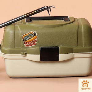 May include: A two-tone green and cream-colored fishing tackle box with a black handle and latch. A sticker on the box reads "Sardine Weenie" with a cartoon image of sardines in a hot dog bun. A fishing rod is placed on top.