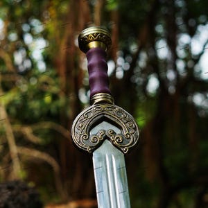 May include: A close-up of a decorative sword with a silver blade and ornate gold and bronze hilt. The handle is wrapped in purple leather. The sword has a detailed, engraved design.