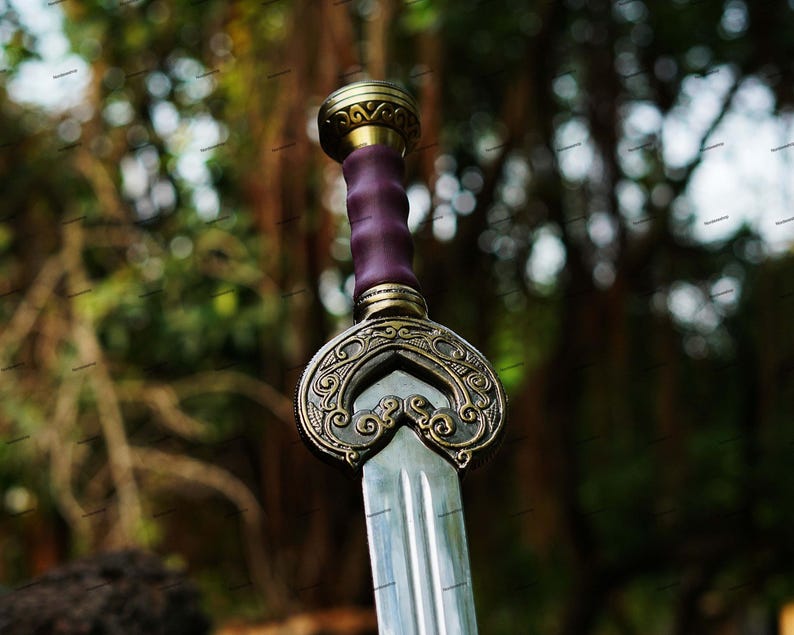 May include: A close-up of a decorative sword with a silver blade and ornate gold and bronze hilt. The handle is wrapped in purple leather. The sword has a detailed, engraved design.
