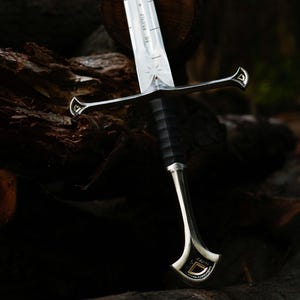 May include: A detailed image of a sword with a silver blade and a black handle. The sword features intricate designs on the hilt and blade, with a gold-colored accent. The sword is resting on a dark, textured wooden surface.