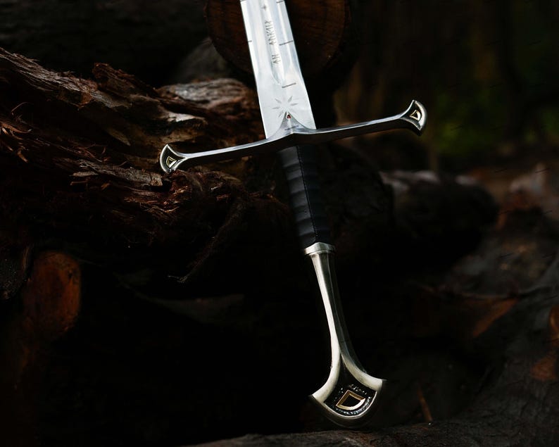 May include: A detailed image of a sword with a silver blade and a black handle. The sword features intricate designs on the hilt and blade, with a gold-colored accent. The sword is resting on a dark, textured wooden surface.