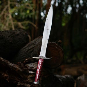 May include: A silver and red dagger with a long, pointed blade. The handle is a deep red with silver accents. The dagger is resting on a weathered log in a forest setting.
