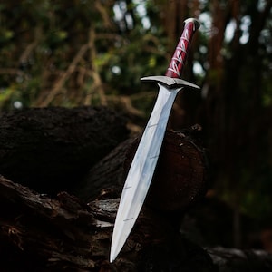 May include: A silver sword with a polished blade and a red and silver handle. The sword is resting on a dark brown log in a forest setting. The handle has a textured design.