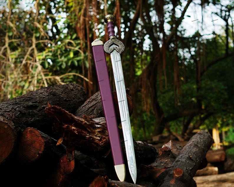 May include: A decorative sword with a silver blade and ornate hilt, resting on a pile of logs. The sword has a purple handle and a matching sheath. The background features lush greenery.