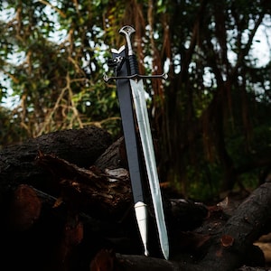 May include: A silver and black sword with a leather-wrapped handle and a silver crossguard is displayed. The sword is sheathed in a black scabbard. The background features a pile of logs and lush green foliage.