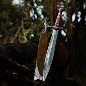 May include: A silver sword with a polished blade and a red and silver handle rests against a brown leather sheath. The sword is displayed on a dark brown log with a blurred forest background.