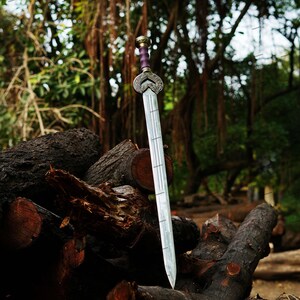 May include: A silver sword with a decorative hilt and a purple handle is displayed against a backdrop of logs and lush greenery. The blade has a series of etched lines. The sword is a fantasy or historical item.