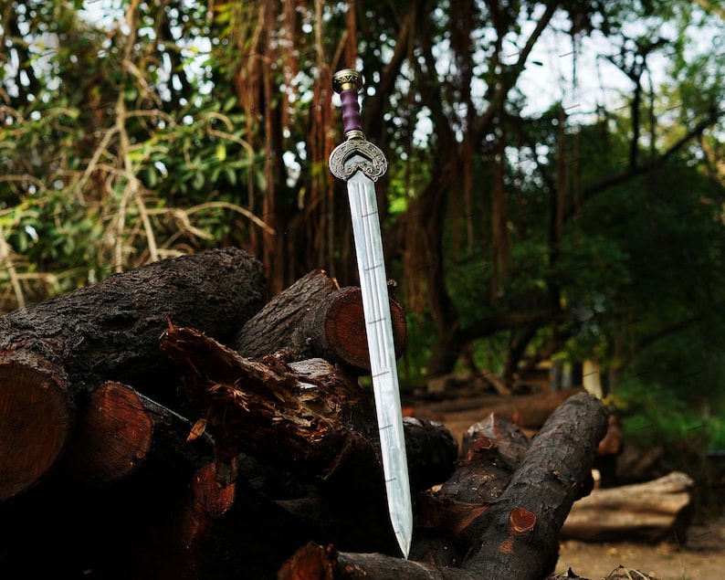May include: A silver sword with a decorative hilt and a purple handle is displayed against a backdrop of logs and lush greenery. The blade has a series of etched lines. The sword is a fantasy or historical item.
