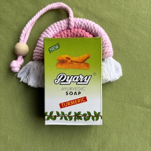 Może przedstawiać: Zielone pudełko mydła ajurwedyjskiego Pyary z kurkumą, na zielonym tle. Na pudełku widnieje nazwa marki i szczegóły produktu. Różowo-biały szydełkowy przedmiot znajduje się za mydłem.