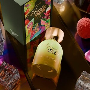 Puede incluir: Un frasco de perfume con tapa dorada y cuerpo degradado amarillo-verde. La botella est&aacute; etiquetada como "Hawaiian Crush Mykonos" en una fuente cursiva. Detr&aacute;s del frasco hay una caja a juego con dise&ntilde;os florales tropicales y el mismo texto. El tema general es la fragancia.
