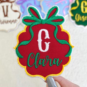 May include: A red embroidered patch shaped like a scalloped ornament with a green bow at the top. The patch features a large white letter C and the name Clara embroidered in green script. The edges are finished with a yellow and white border.