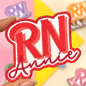 May include: Embroidered patch with the letters "RN" in red, with the name "Annie" in cursive script. The patch has a white background and a red border. Other patches are visible in the background.