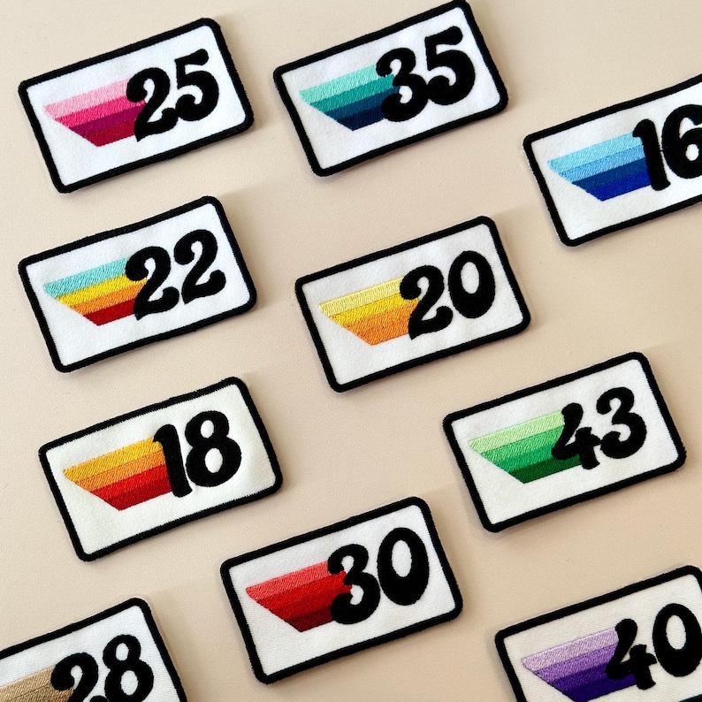 May include: A selection of embroidered patches with white backgrounds and black borders. Each patch displays a number in bold black font and a colourful, angled stripe design. The numbers range from 16 to 43, with various colour combinations.