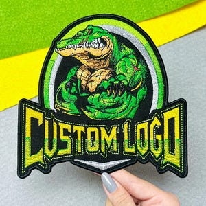 May include: An embroidered patch featuring a muscular green alligator with sharp teeth, positioned within a circular frame. Below the alligator, the text CUSTOM LOGO is stitched in bold yellow letters with a black outline on a black background.