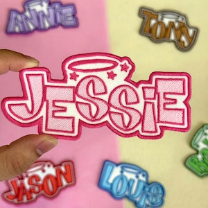May include: Embroidered patch with the name "Jessie" in pink, outlined in a darker pink, with a halo and stars design. Other name patches are visible in the background.