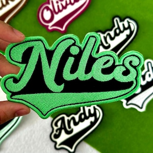 May include: Green embroidered patch with the name "Niles" in a black, stylized font. The patch has a white border and is surrounded by other patches with names. The background is a solid green color.