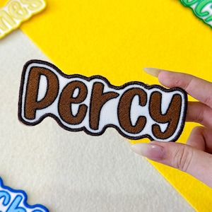May include: An embroidered patch featuring the name "Percy" in brown lettering with a white outline and a brown border. The patch is set against a yellow and white background. Other patches are visible.