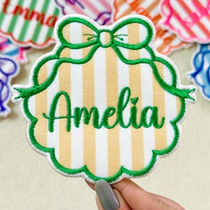 May include: A scalloped, embroidered patch featuring a light yellow and white vertical striped background. The name Amelia is stitched in green cursive script across the center, topped with a green embroidered bow and decorative border.