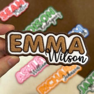 May include: Embroidered patch featuring the name "Emma Wilson" in brown thread on a white background, with a brown border. Other name patches in various colors are visible in the background.