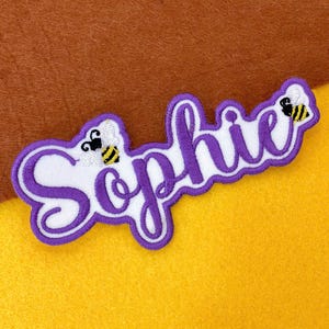 May include: Embroidered patch spelling "Sophie" in a cursive font, in purple with a white background and a purple border. Two cartoon bees are on the patch. The patch is on a yellow and brown background.