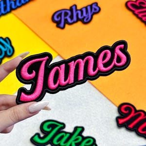 May include: Embroidered name patches in various colors. The patch held in the hand says "James" in pink script with a black border. Other patches with names are visible in the background, including "Rhys" and "Jake". The patches are on colorful backgrounds.