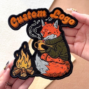 May include: Embroidered patch featuring a fox in a green sweater, holding a mug, with a campfire and the words "Custom Logo". The fox is orange and white, with a black outline. The campfire has yellow and orange flames. The text is in orange.