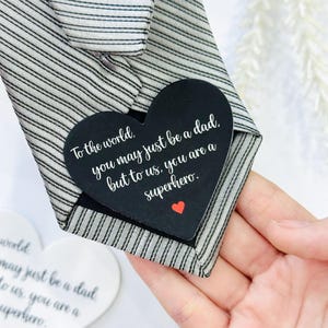 May include: A striped tie with a black heart-shaped tag. The tag has the text "To the world, you may just be a dad, but to us, you are a superhero." and a small red heart.