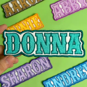 May include: Embroidered patch with the name "DONNA" in teal letters with a white border. The patch is held against a green background, with other name patches visible. The letters have a decorative, vintage style.