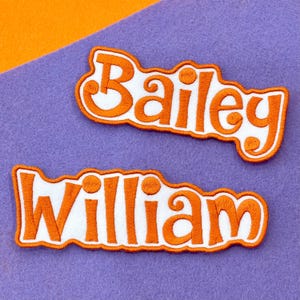 May include: Two embroidered patches with white lettering and orange outlines. The top patch reads "Bailey" and the bottom patch reads "William". The patches are on a purple felt background with an orange felt accent.