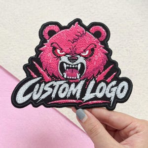 May include: An embroidered patch featuring a stylized pink bear head with an aggressive expression, sharp teeth, and red eyes. Below the bear, the text CUSTOM LOGO is stitched in white with a black outline on a black background.