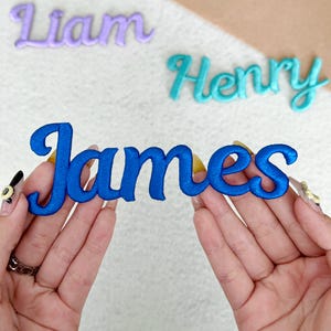 May include: Close-up of embroidered name patches. The name "James" is in bright blue, held by hands. Other names, "Liam" in purple and "Henry" in teal, are visible in the background. The patches have a cursive font and a stitched border.
