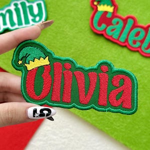 May include: Embroidered patch with the name "Olivia" in red letters, outlined in green. The patch features a green elf hat with a gold crown. The background is a light green felt.
