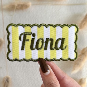 May include: A rectangular embroidered patch with a scalloped edge featuring a yellow and white vertical striped pattern. The name Fiona is stitched in dark olive green cursive lettering across the center of the patch with a matching green border.