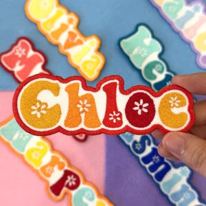 May include: Embroidered name patches in various colors, including orange, red, yellow, and blue. The patches feature names like "Chloe" and "Olivia" with floral accents. The patches are on a blue and purple background.