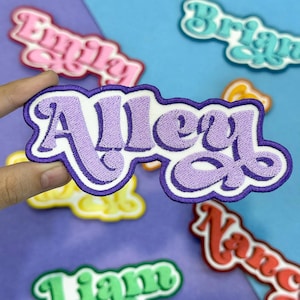 May include: A selection of embroidered name patches in different colours. The central patch reads "Alley" in purple thread, with a white border and a purple outline. Other patches with names such as "Emily", "Brian", and "Nancy" are visible.