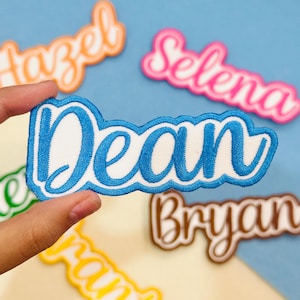May include: A blue embroidered name patch featuring the name Dean in a cursive script font with a white background and a blue border. The patch has a scalloped edge design and is displayed alongside other similar name patches in various colors.