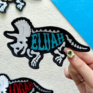 May include: An embroidered iron-on patch shaped like a triceratops dinosaur skeleton. The patch features a black background with white skeletal details and the name ELIJAH stitched in bright teal blue thread across the center of the body.