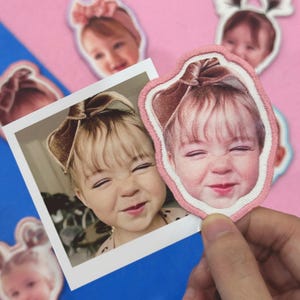 May include: A close-up of a smiling baby's face on a pink embroidered patch with a white border. The baby has a brown bow in their hair. A polaroid photo of the same baby is also visible, along with other similar patches and stickers.