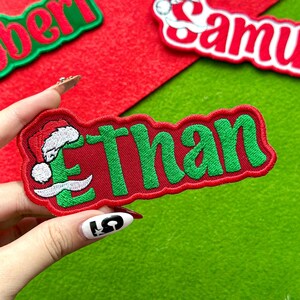 May include: A red embroidered patch with the name "Ethan" in green letters. The letter "E" features a Santa hat and mustache design. The patch is on a green and red felt background.