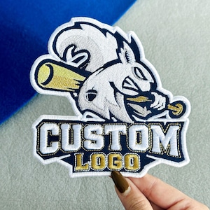 May include: An embroidered patch featuring a stylized white squirrel mascot holding a yellow baseball bat. Below the mascot, a dark blue banner with a white border contains the text CUSTOM LOGO in bold white letters with a gold-colored outline.
