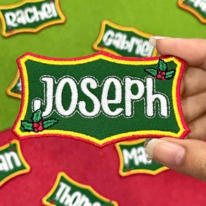 May include: A green embroidered patch with the name "Joseph" in white, framed by a yellow and red border. The patch features holly berry accents and is held against a red and green background with other name patches.