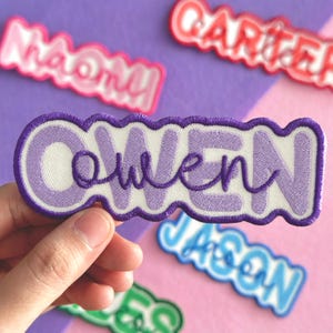 May include: Embroidered patch with the name "Owen" in purple script lettering. The patch has a white background and a wavy purple border. Other name patches are visible in the background, including "Naomi" and "Carter".