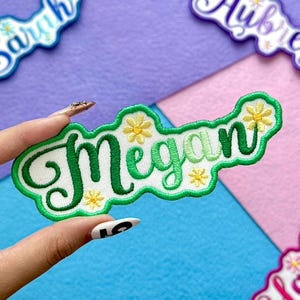May include: Embroidered patch with the name "Megan" in green script, outlined in green, and adorned with yellow flower accents. The patch is on a white background and is surrounded by other patches on a purple and pink background.