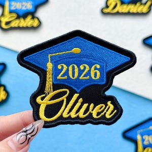 May include: Embroidered patch featuring a blue graduation cap with the year 2026 and the name "Oliver" in gold lettering. The patch has a black border. Other patches with names and the year 2026 are visible in the background.