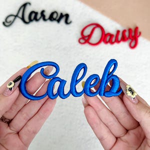 May include: Embroidered name patches in black, red, and blue. The blue patch displays the name "Caleb" in a cursive font, held by hands. The names "Aaron" and "Daisy" are also visible. These patches are suitable for personalizing clothing or accessories.