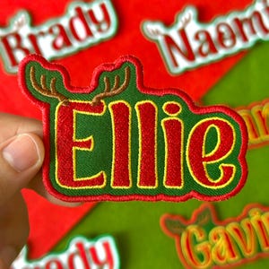 May include: Embroidered patch with the name "Ellie" in red letters outlined in yellow, set against a green background. The patch features a reindeer antler design above the name and is surrounded by a green border. Other name patches are visible in the background.