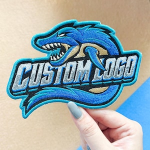 May include: An embroidered patch featuring a stylized blue shark head with sharp teeth and gold accents. The text CUSTOM LOGO is stitched in silver and black across the center. The design is outlined in bright blue thread with a black border.