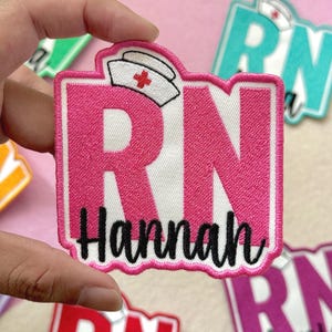 May include: A pink and white embroidered patch with the letters "RN" and the name "Hannah" in black script. The patch features a nurse's cap with a red cross. The patch is rectangular with a white border.