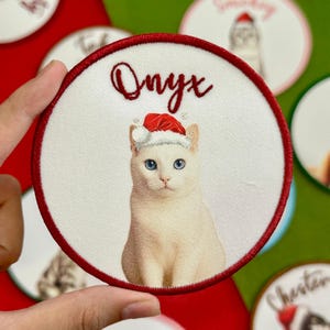 May include: A round, white patch with a red border featuring a white cat wearing a Santa hat. The name "Onyx" is written in red script above the cat. Other similar patches are visible in the background.