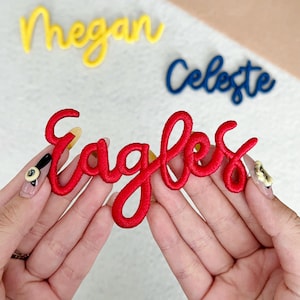 May include: A red embroidered "Eagles" script is held in hands, with yellow "Megan" and blue "Celeste" script in the background. The cursive script is ideal for customising clothing or craft projects.